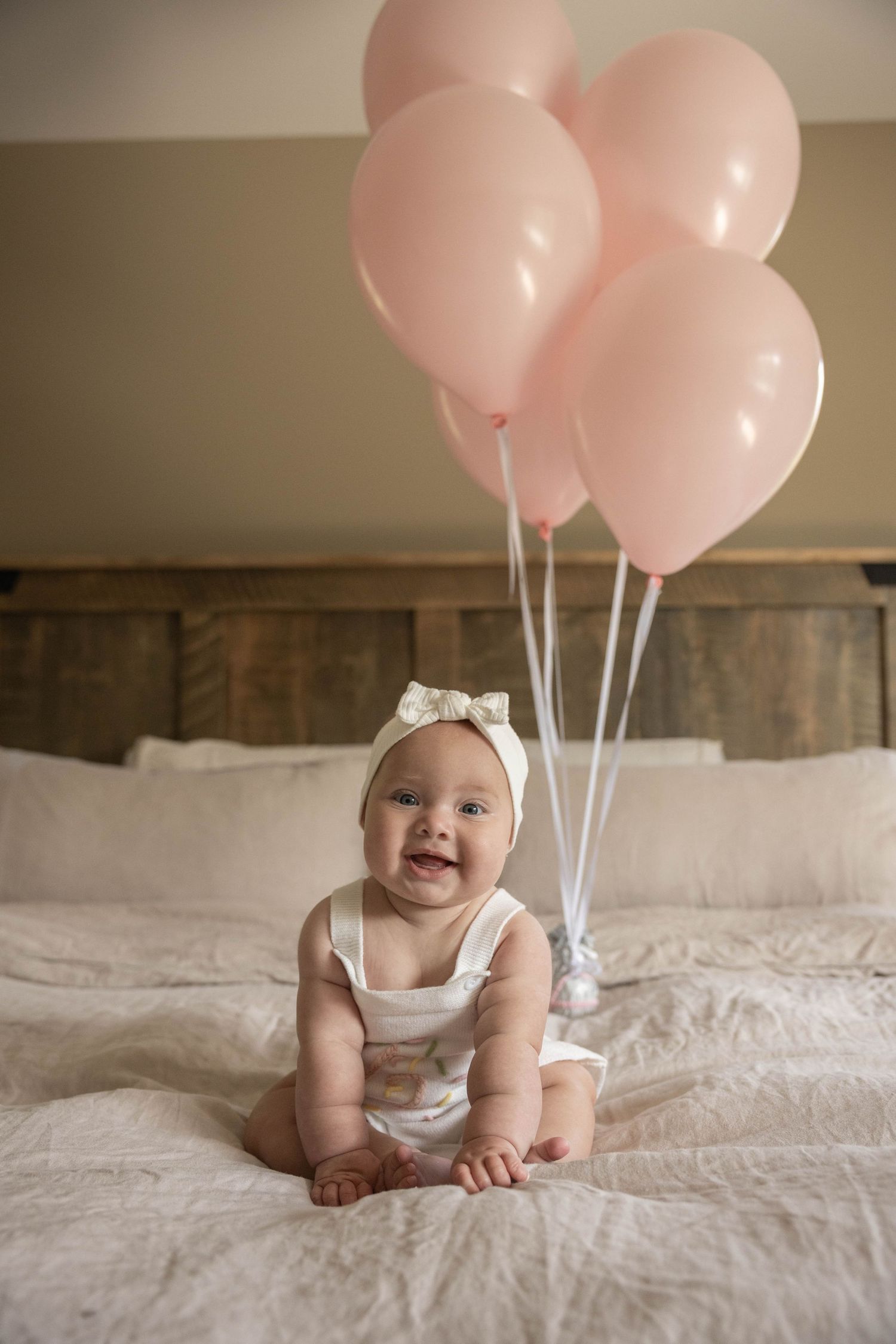 Smiling baby girl in white romper and bow headband sitting on bed with pink balloons.