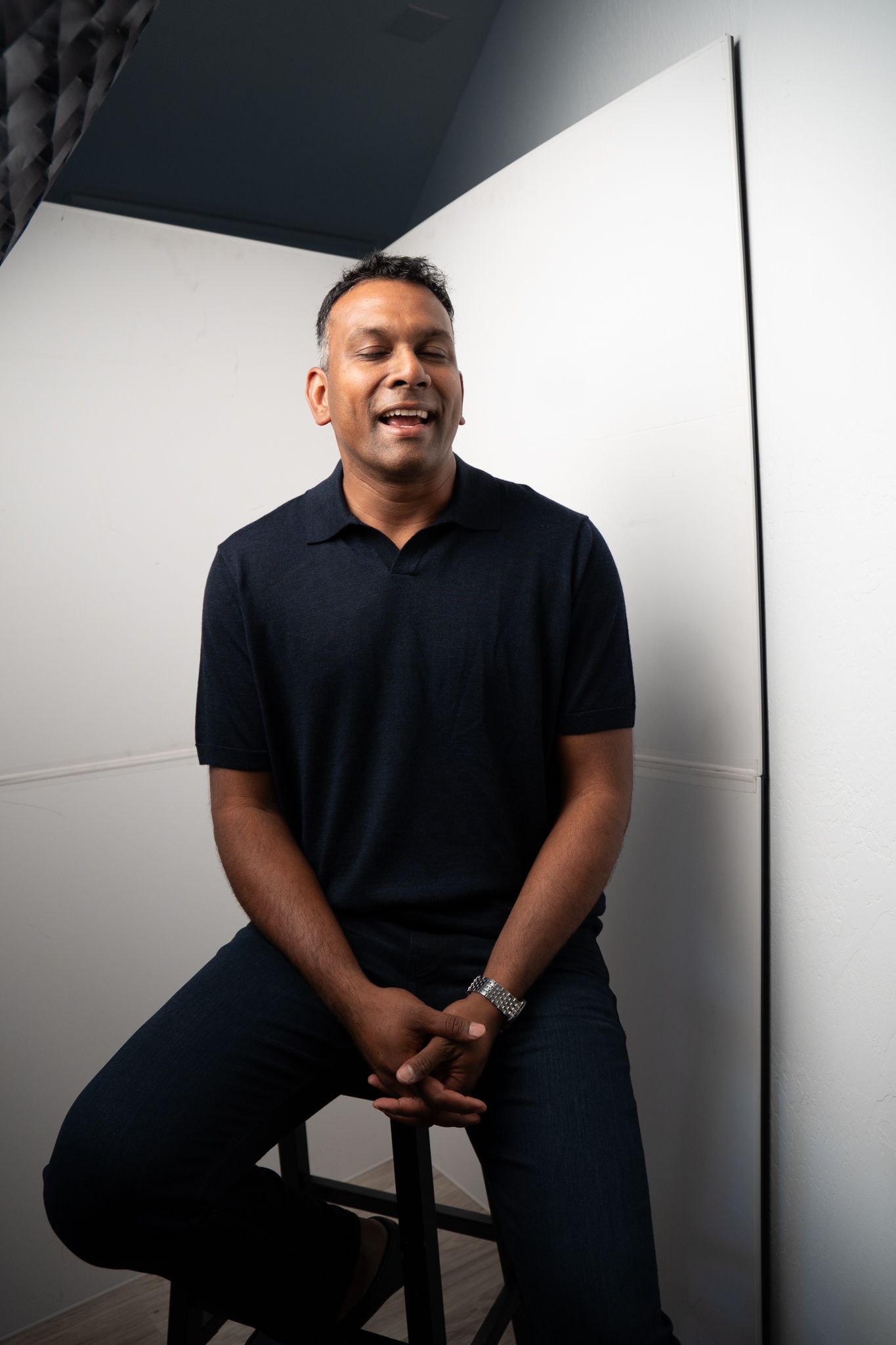 Smiling man in black polo shirt sitting on stool against white wall in professional photo studio setting.