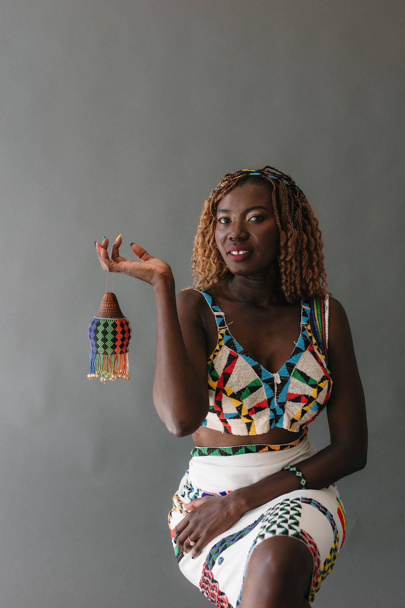 A woman in colorful African print crop top and skirt holds a beaded decorative item against a gray background.
