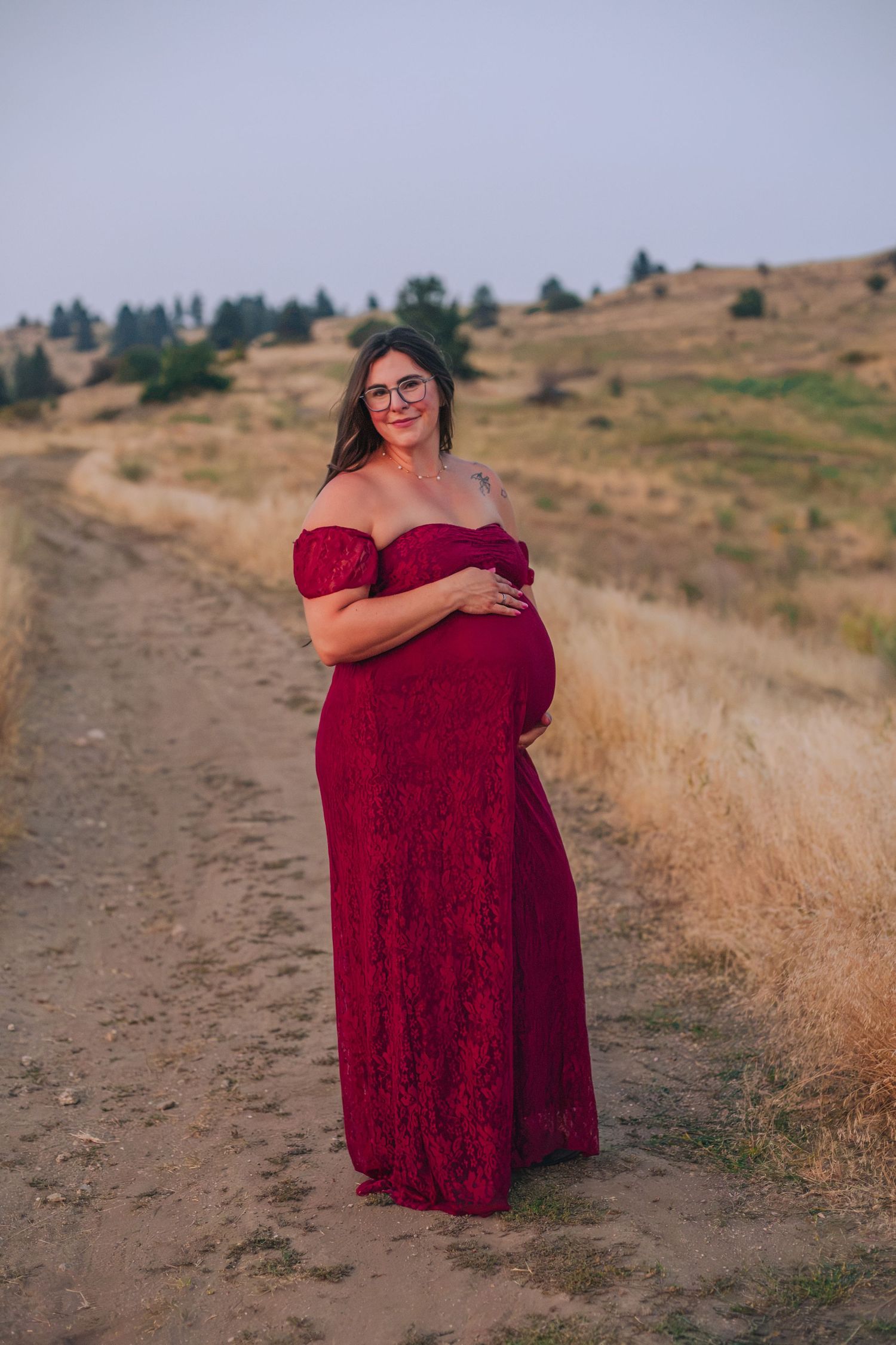 Pregnant woman in red lace off-shoulder gown poses on rural dirt path surrounded by golden fields.