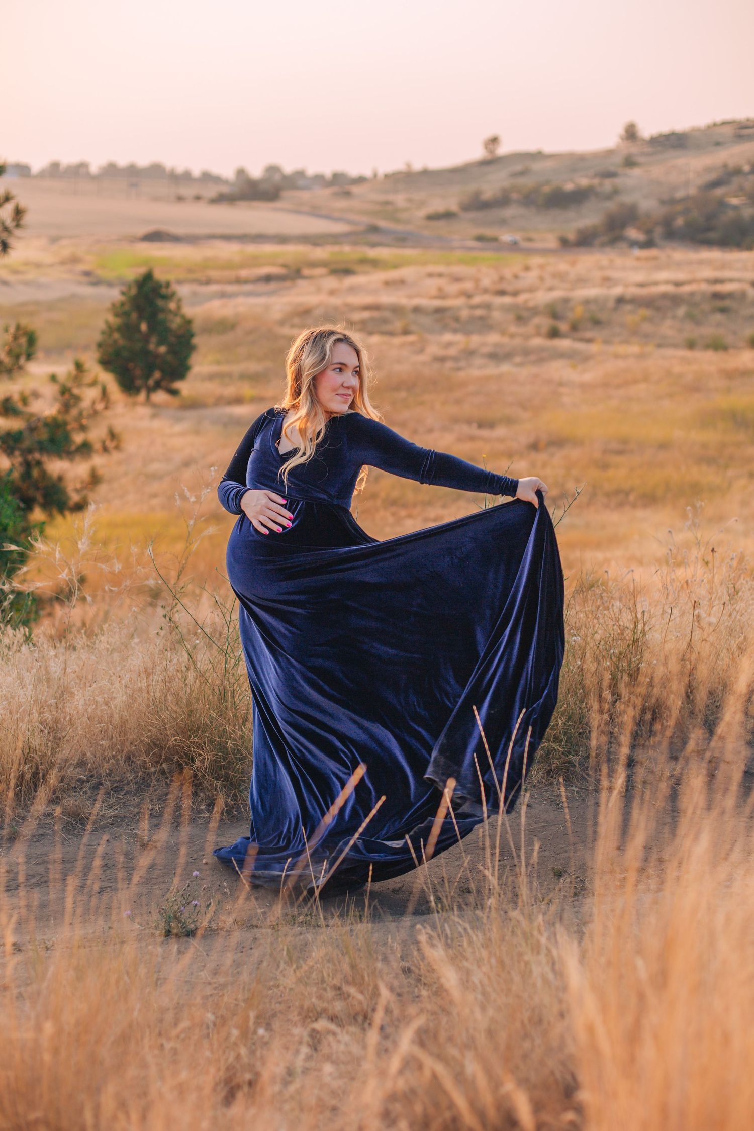 Pregnant woman in blue velvet gown posing in golden field at sunset.