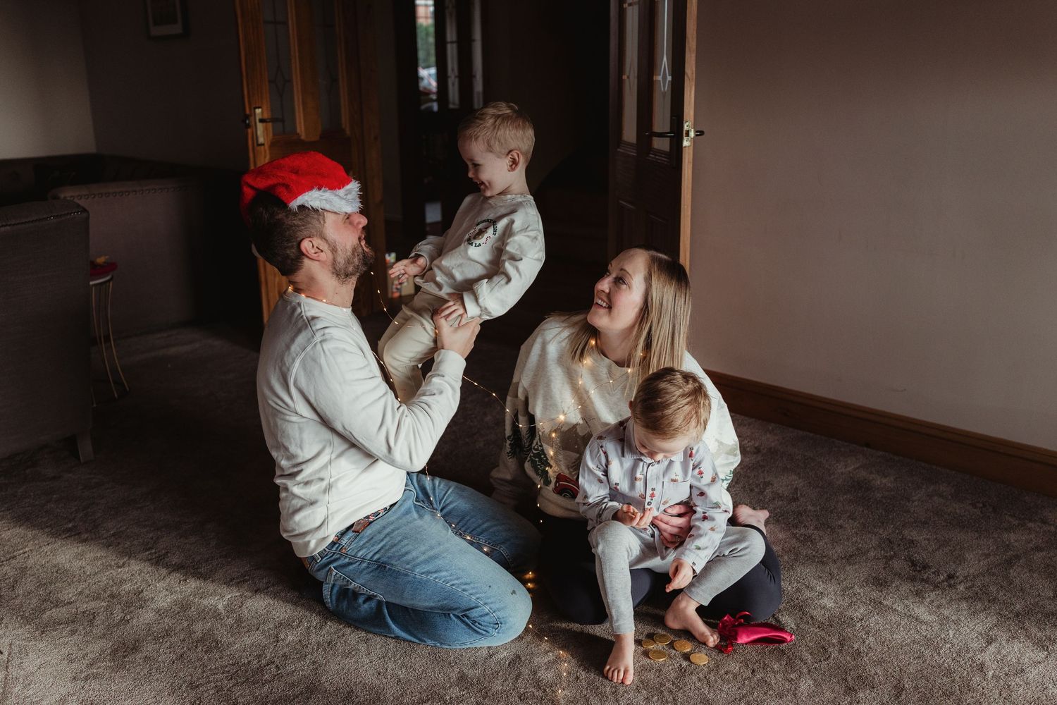 Family sitting on carpet floor celebrating Christmas, dad wearing Santa hat, mom holding baby girl, toddler standing nearby.