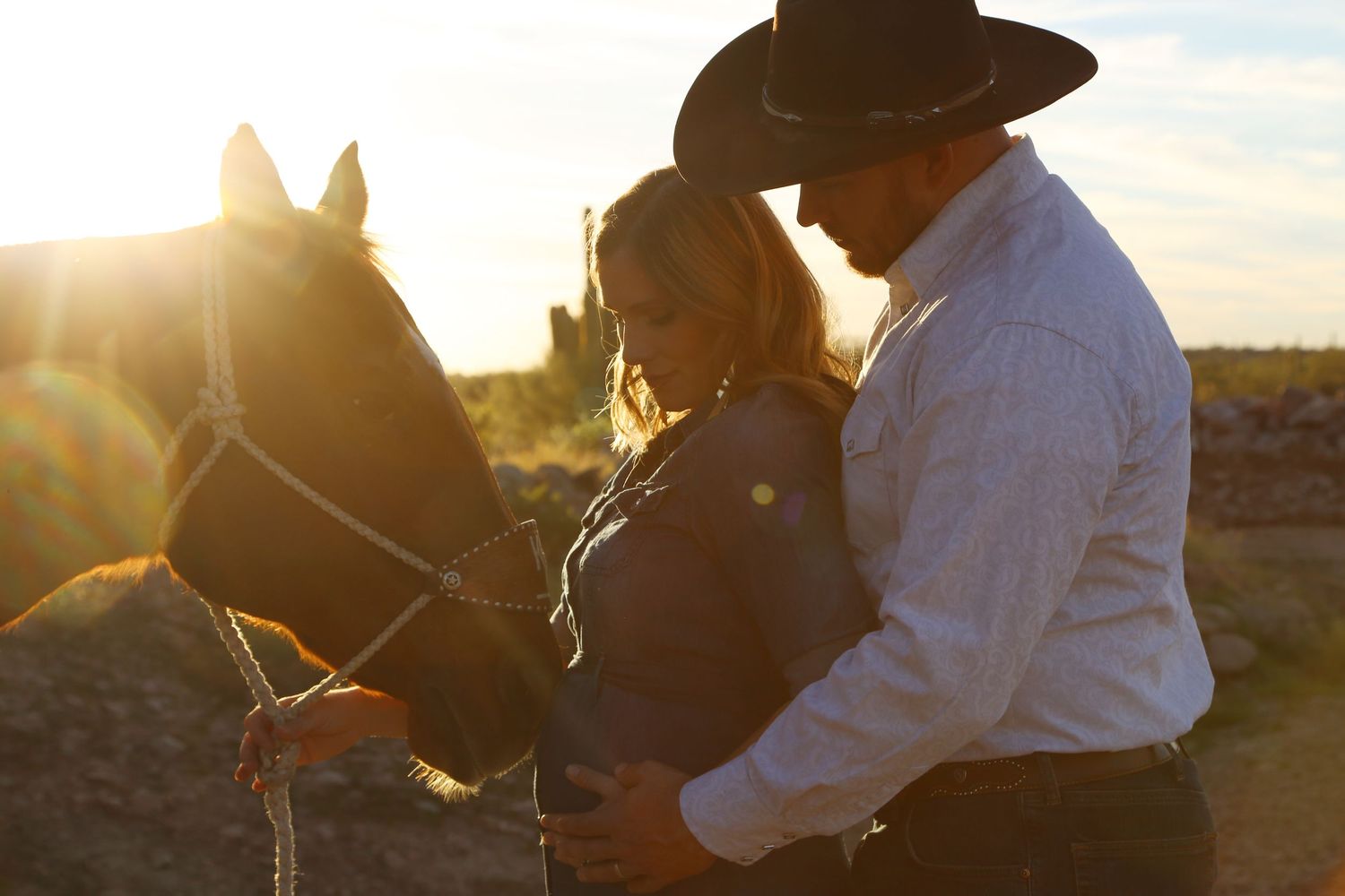 Cowboy in a black hat and a woman embrace beside a horse in the warm glow of a desert sunset.