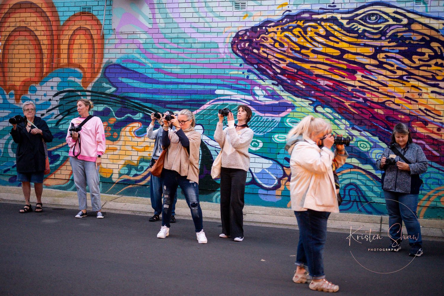 Group of photographers capturing colorful fish mural street art on brick wall outdoors.