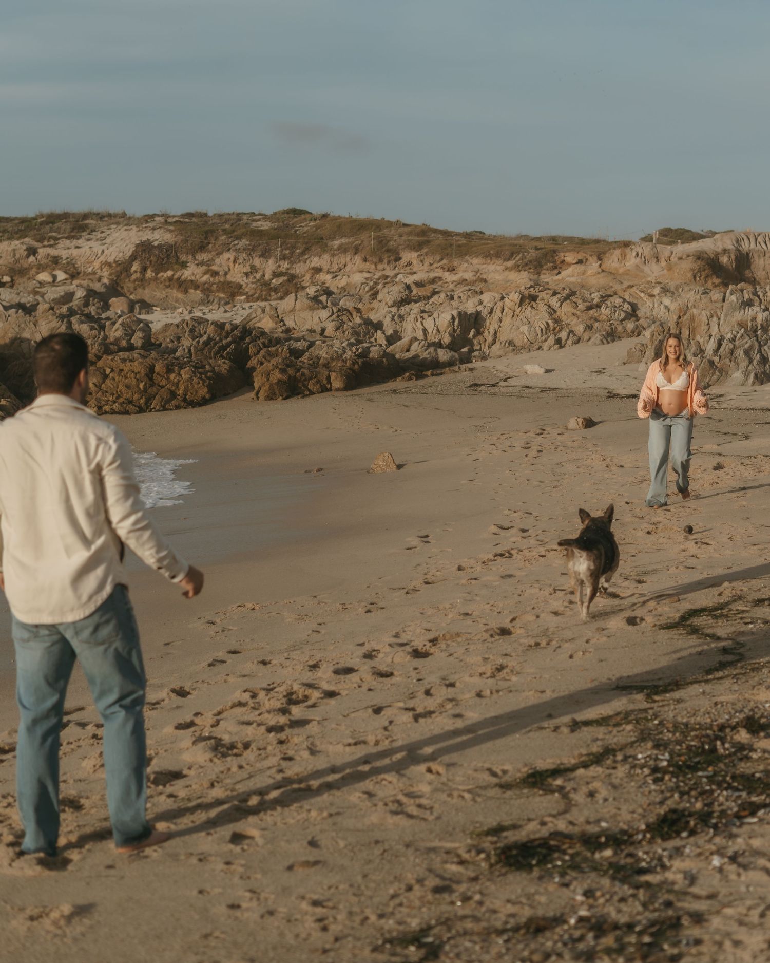 Two people and a dog play on a sandy beach with rocky cliffs in the background during golden hour.