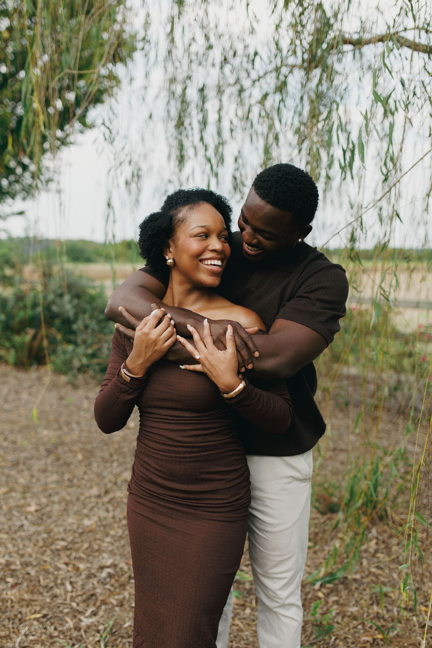 A joyful Black couple embraces outdoors, woman in brown dress laughing as man hugs her from behind near willow tree.