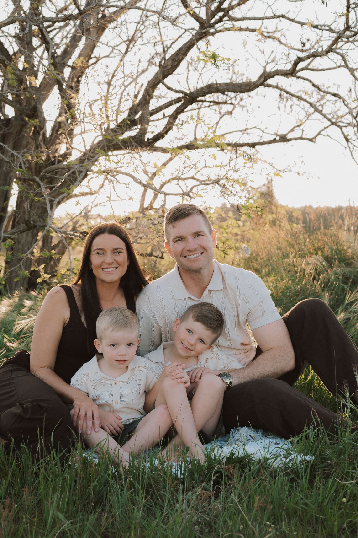 Family of four smiling outdoors, sitting in green grass under a tree in warm golden sunlight.