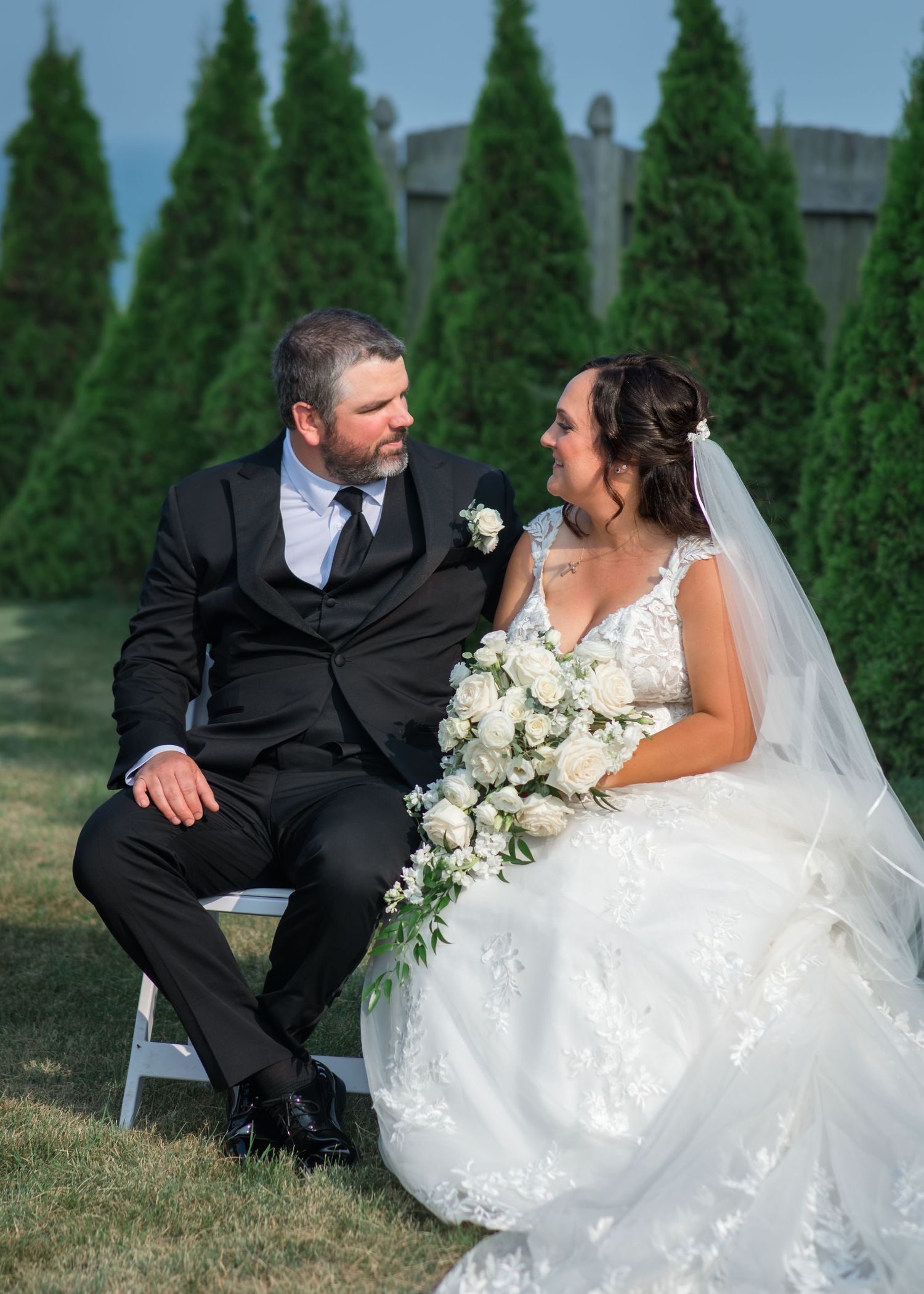 A bride and groom sit together on white chairs outdoors, sharing a loving glance, surrounded by tall green trees.