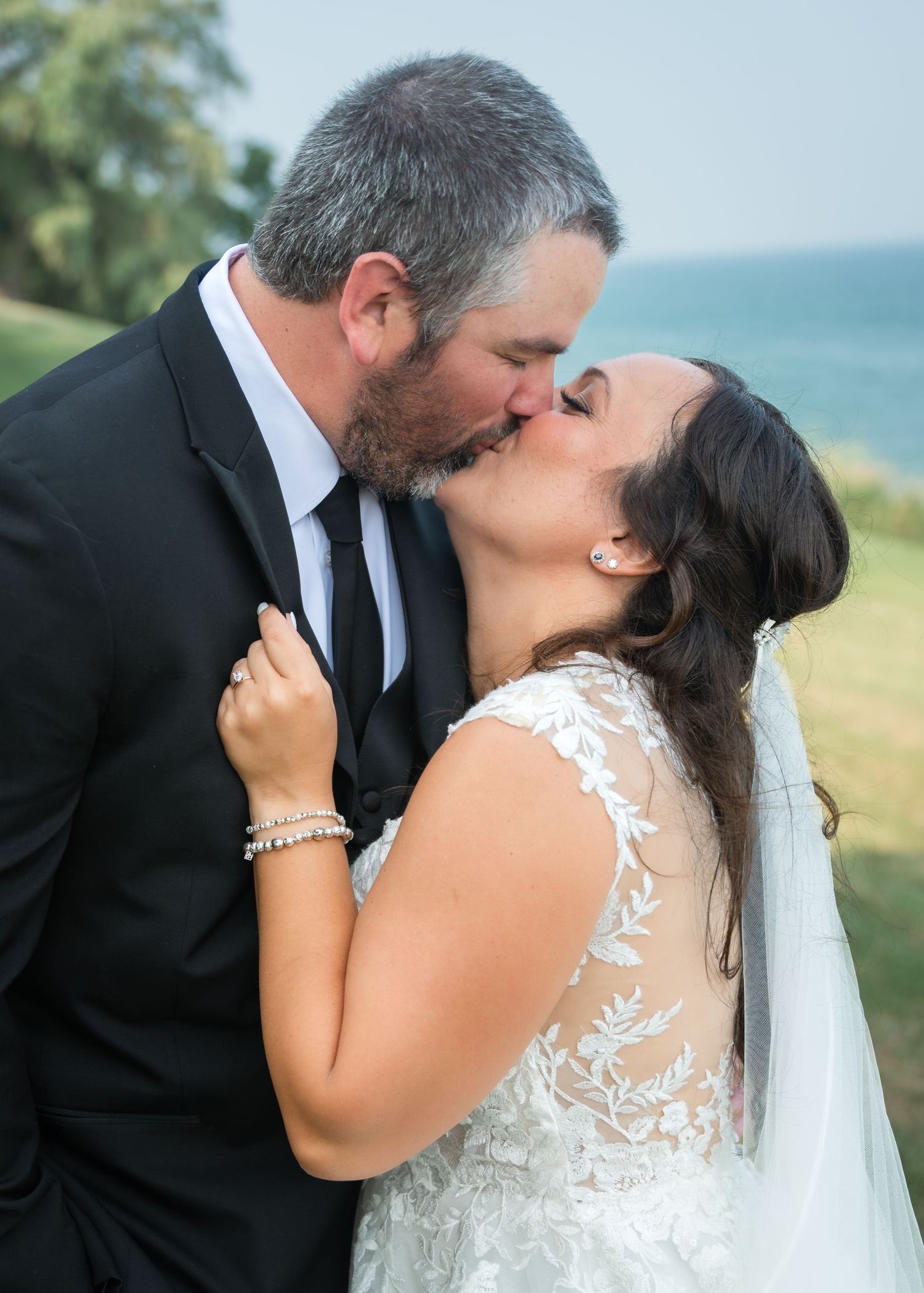 A bride and groom share a romantic kiss outdoors, with the bride wearing a lace wedding gown and veil.
