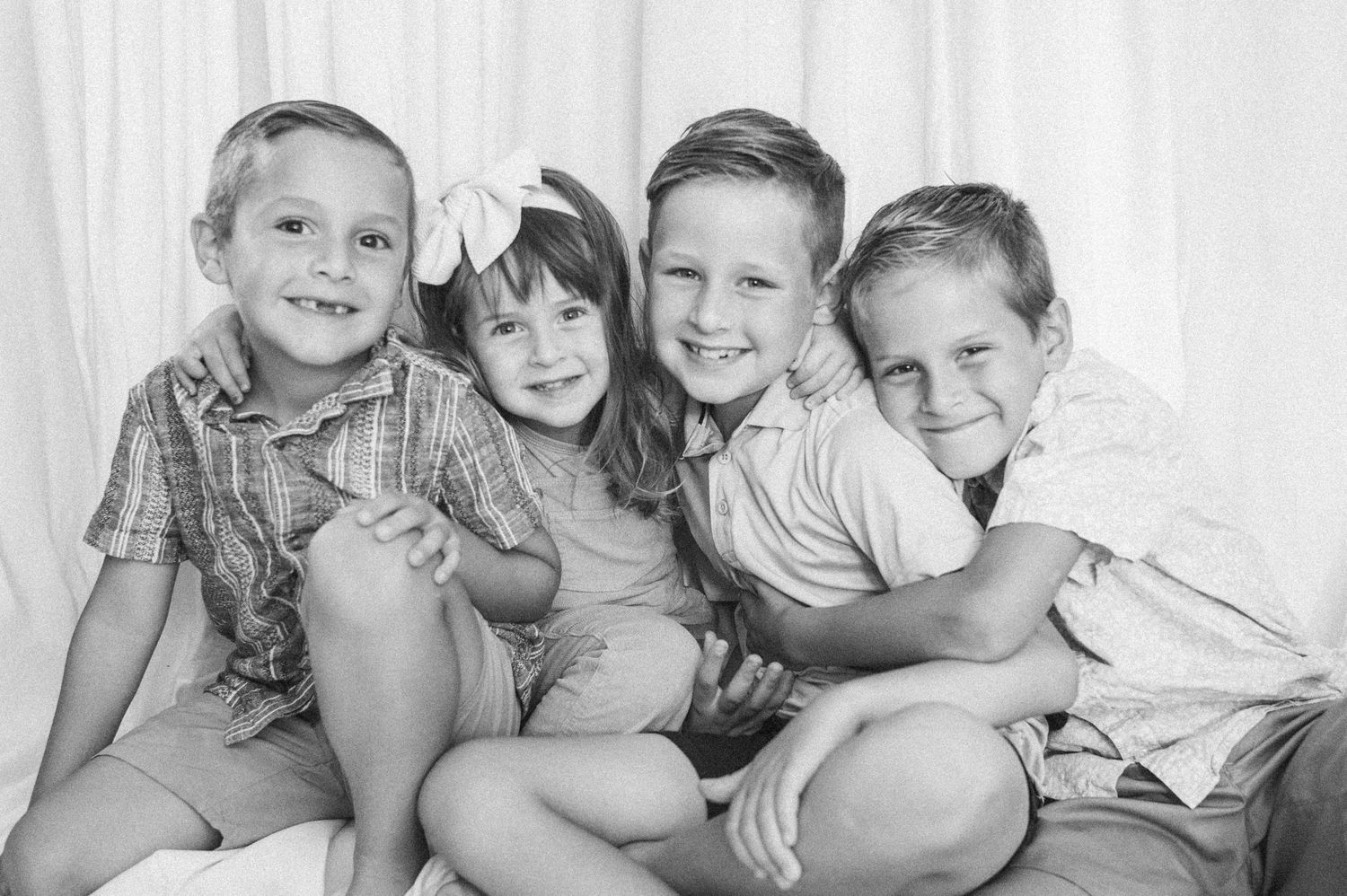 Black and white portrait of four smiling siblings hugging together on white fabric backdrop.