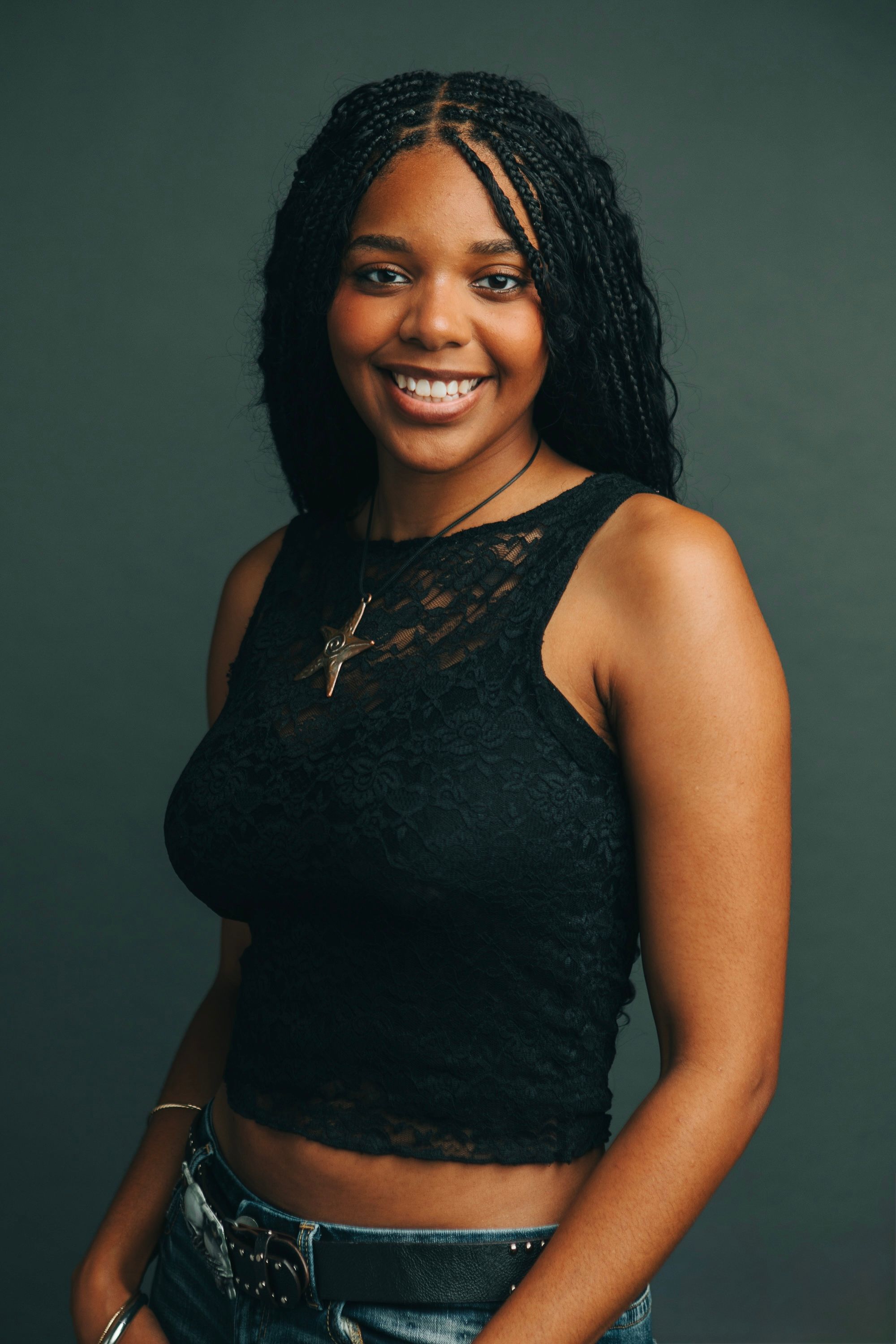 Young Black woman with box braids smiling, wearing black lace crop top and cross necklace against dark background.