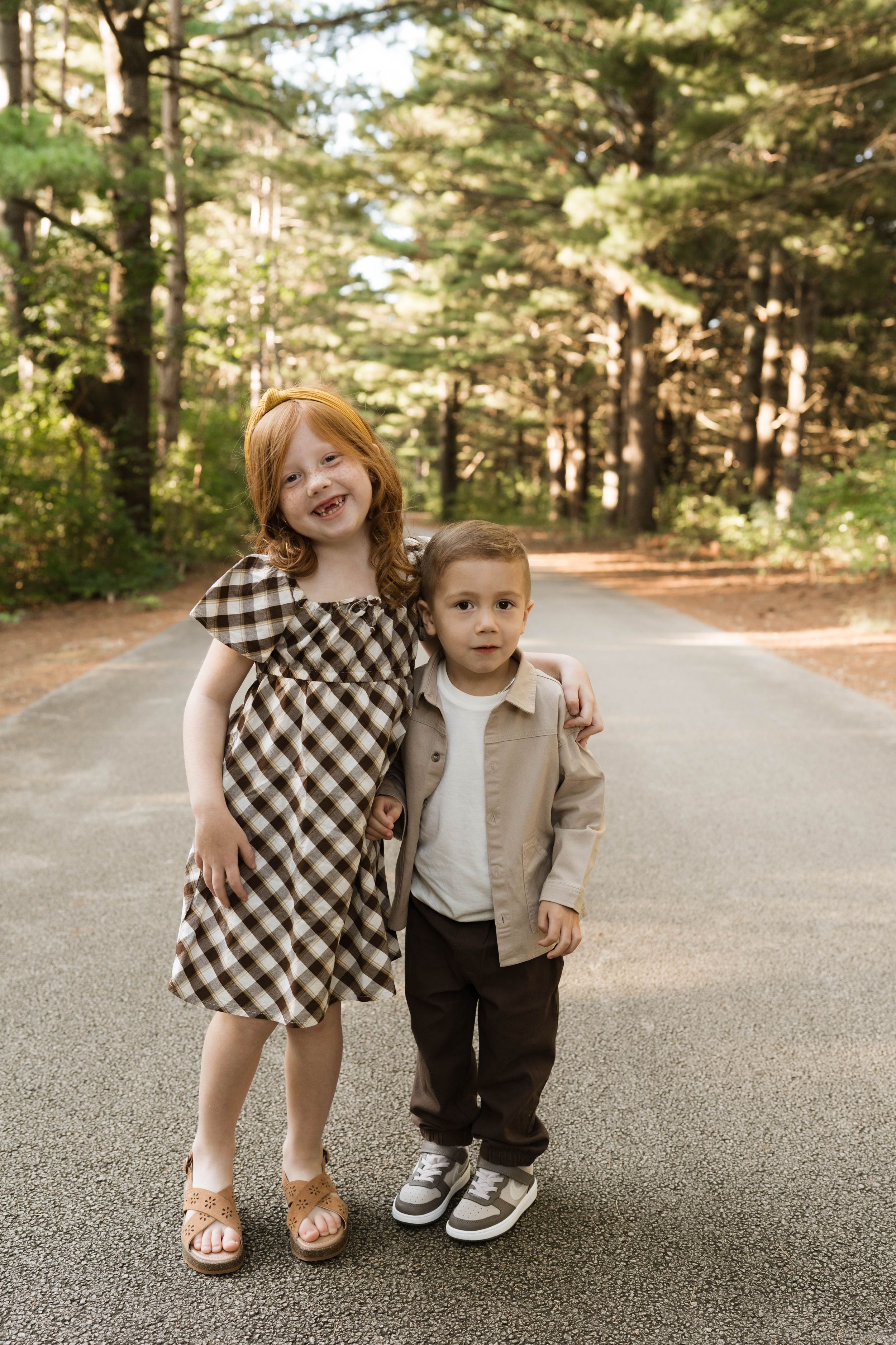 Two young children pose on a tree-lined path, girl in plaid dress with red hair hugging younger boy in tan jacket.