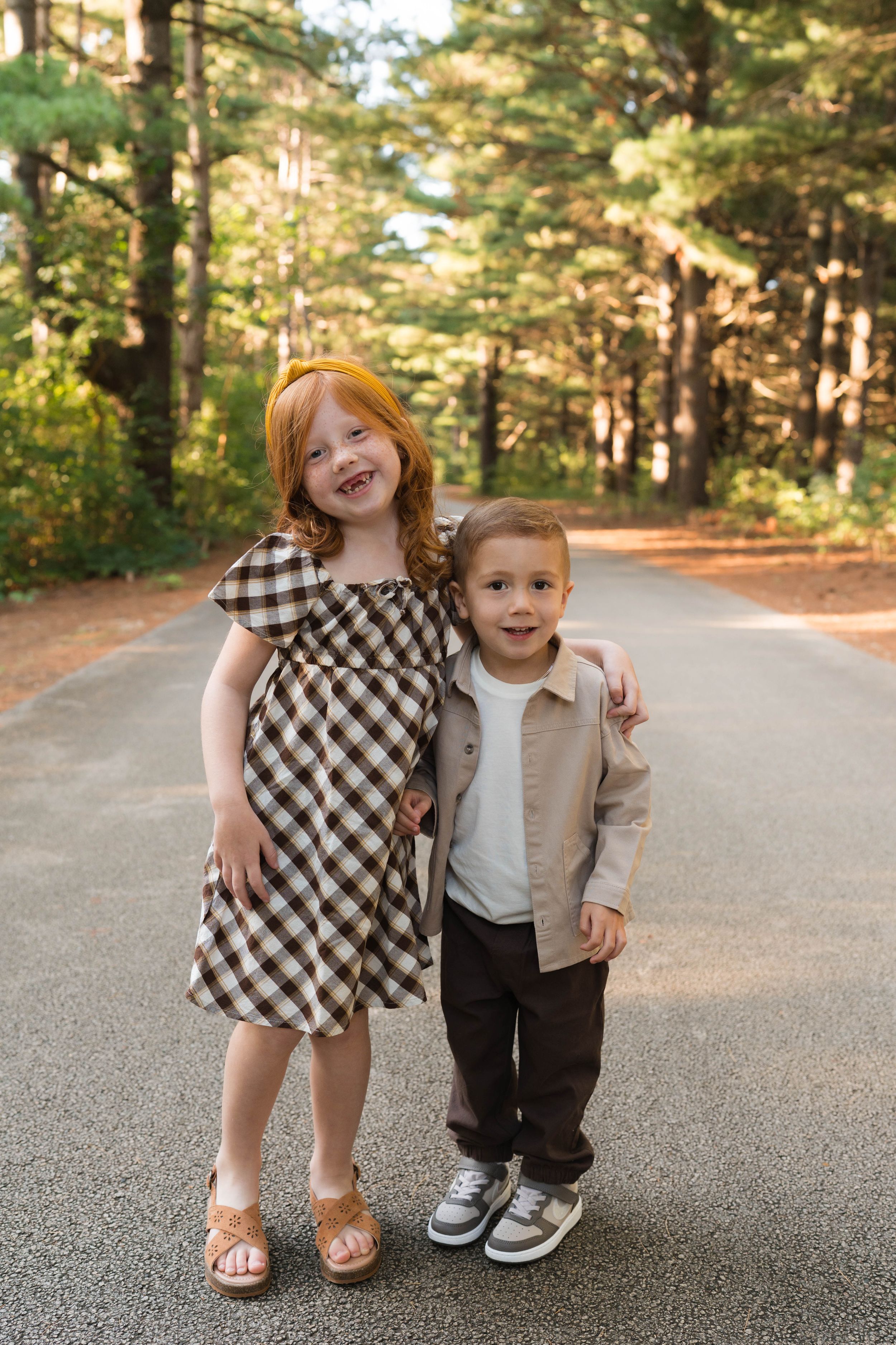 Two young children, a red-haired girl in plaid dress and a boy in beige jacket, pose together on a tree-lined path.
