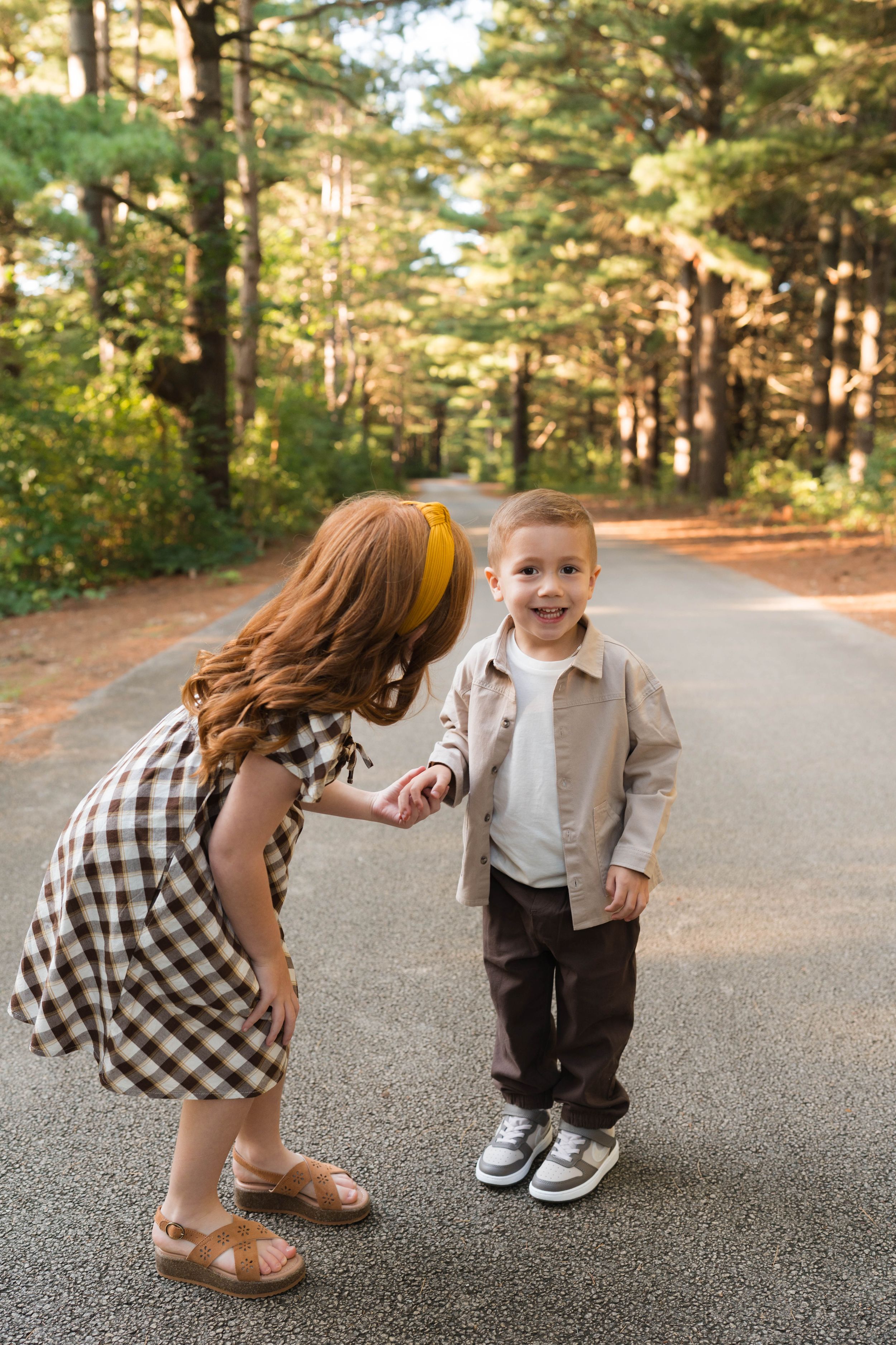 A young girl in a plaid dress holds hands with a smiling toddler boy on a tree-lined path.