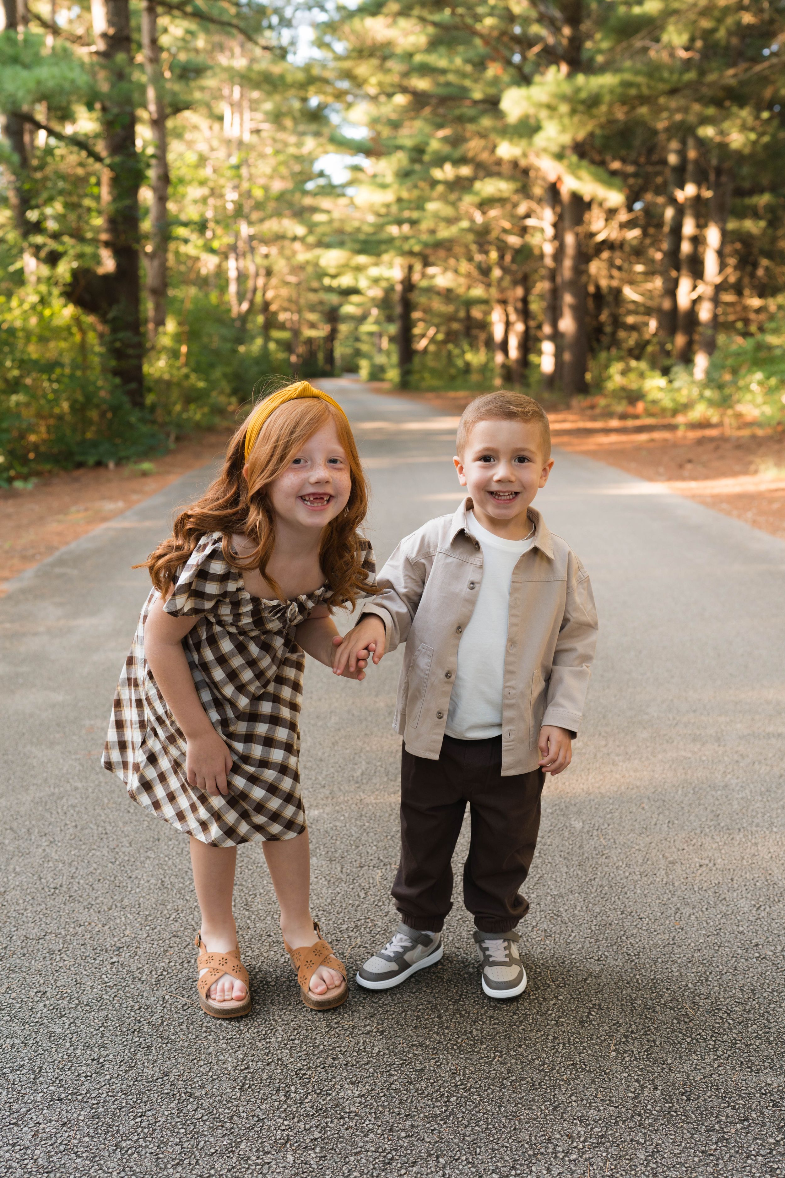 Two young children holding hands on a tree-lined road, girl in plaid dress and boy in beige shirt.