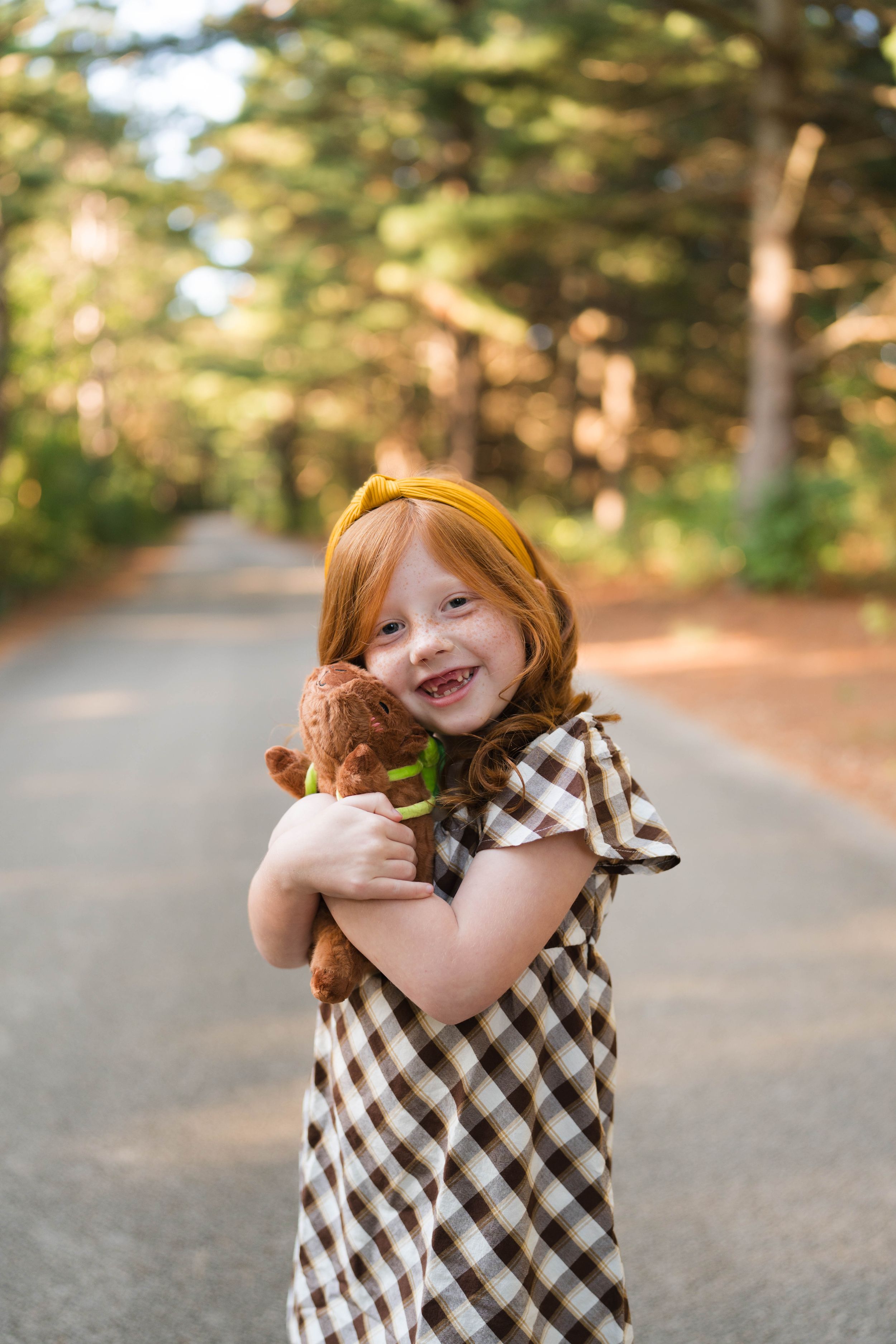 A smiling redhead girl in a plaid dress hugs a brown teddy bear on a tree-lined path.