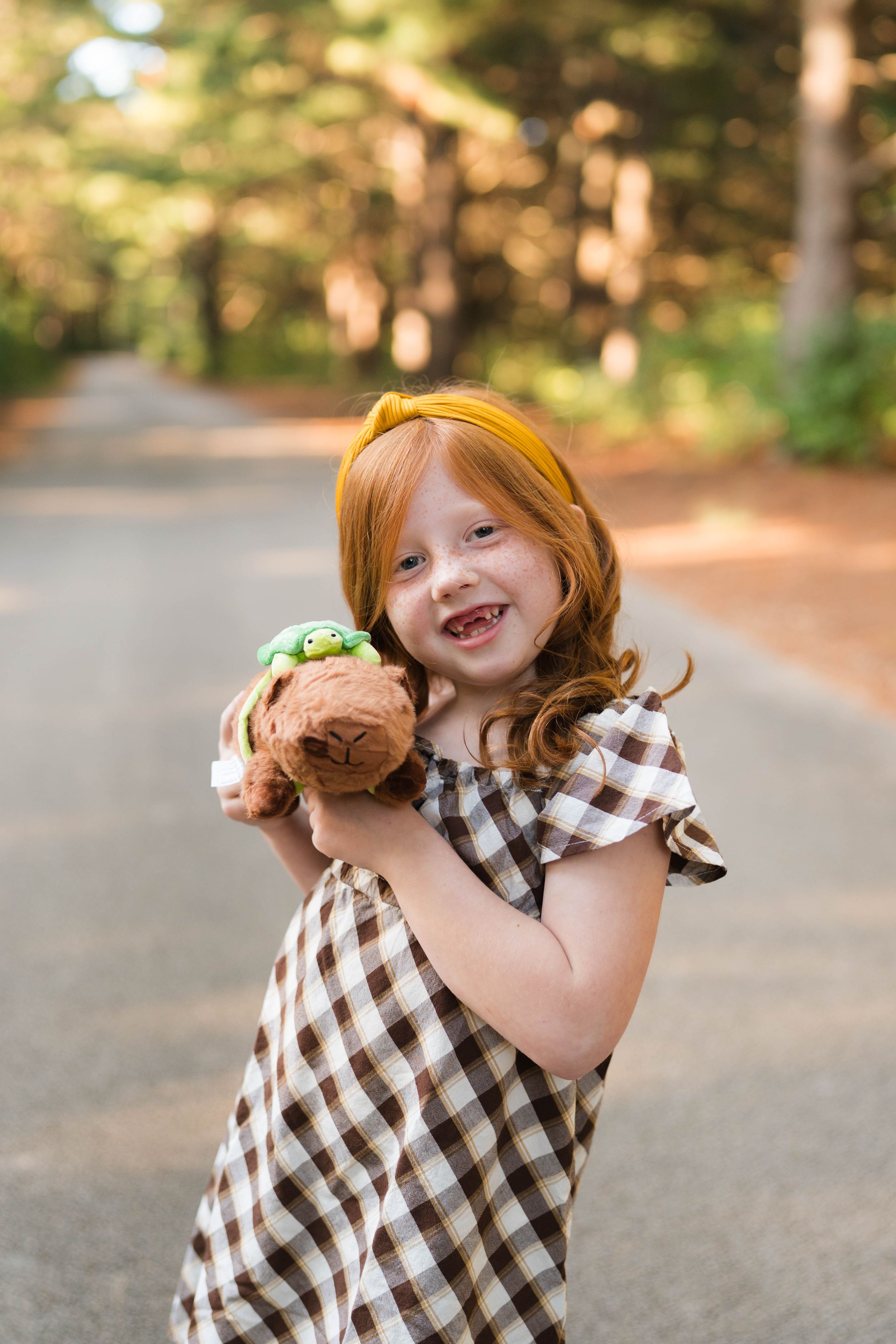 Smiling red-haired girl in plaid dress holds a stuffed turtle toy on a sunlit forest path.