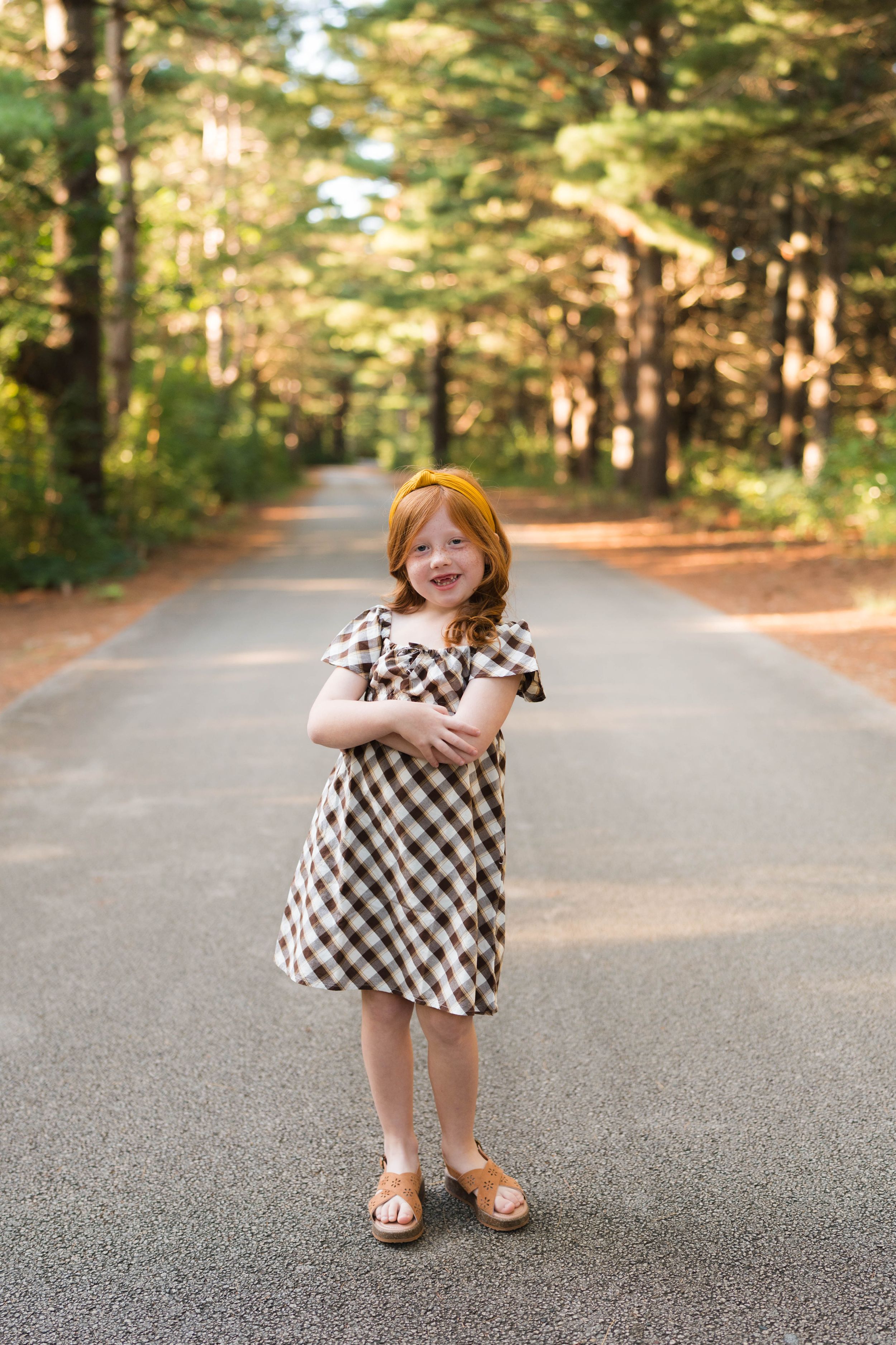 A smiling red-haired girl in a plaid dress stands on a tree-lined path in a sunny forest.