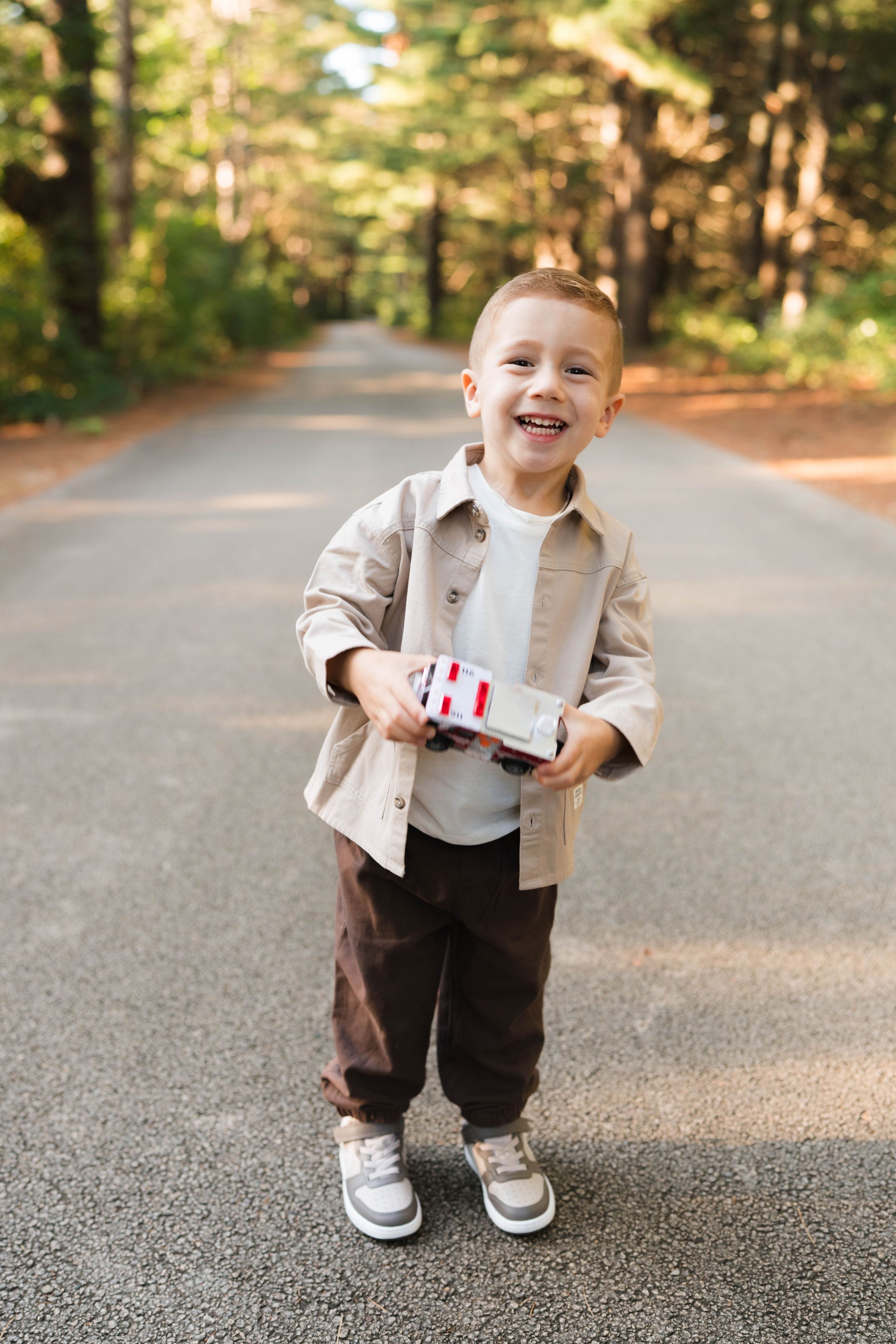 A happy toddler boy in a beige shirt holds a toy car while standing on a sunny forest path.