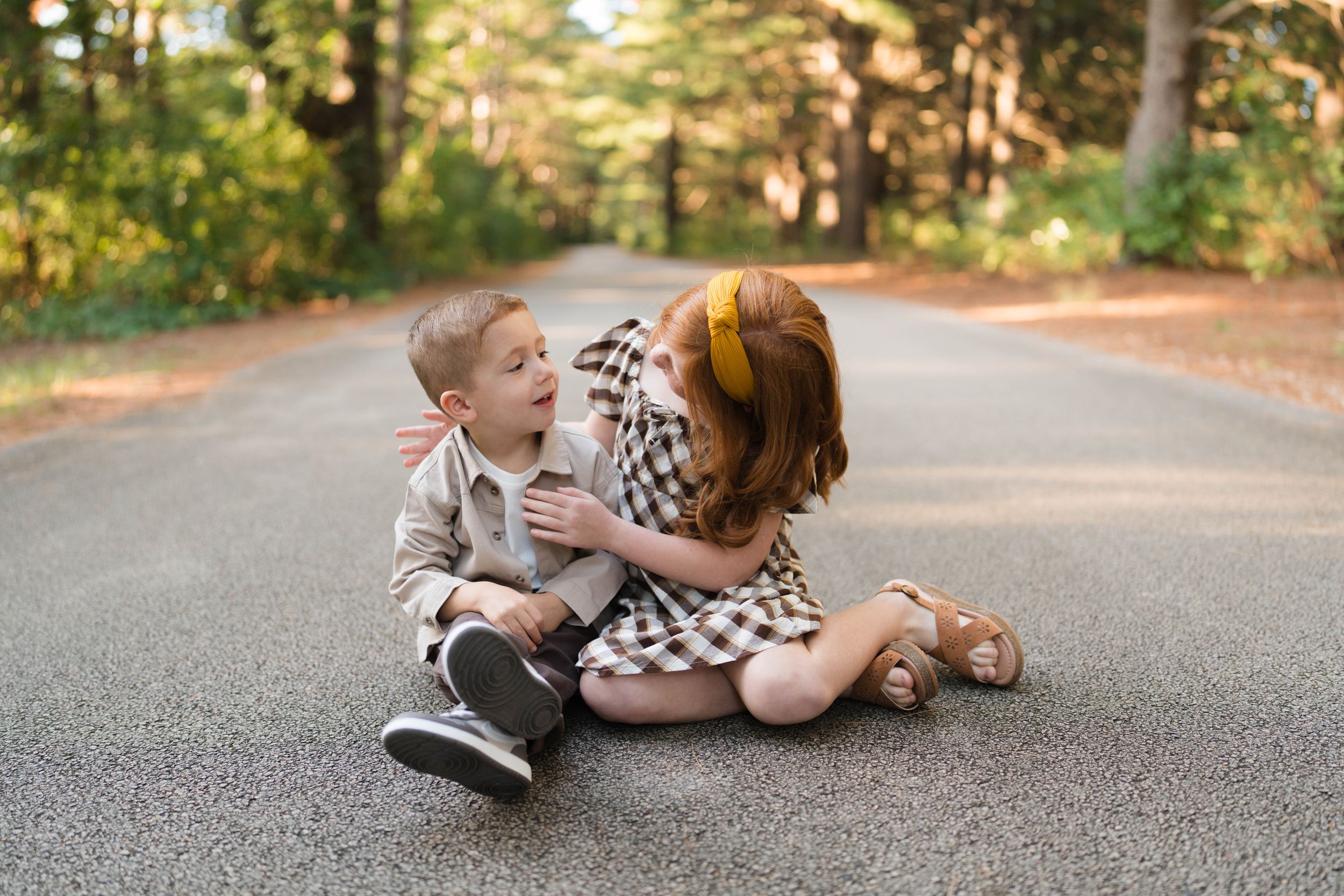Two young children sit together on a tree-lined road, sharing a sweet moment in autumn outdoor portrait.