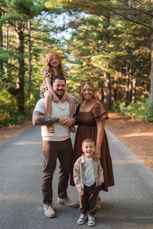 Family of four poses on a tree-lined path, parents with two young redhead children, smiling outdoors.