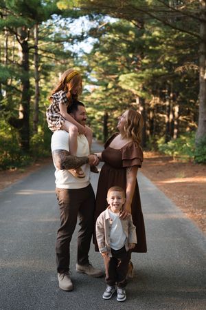 Family of four posing on a tree-lined path, parents smiling at each other while young children laugh joyfully.
