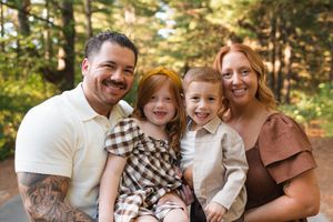 Smiling family of four poses outdoors among trees, featuring dad with tattoos, mom, and two young redheaded children.