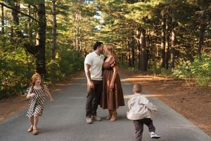 Family of four on a tree-lined path, parents kissing while children play around them in a sunlit forest.