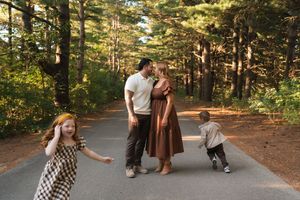 Family of four on a forest path, parents kissing while two young children play around them in golden sunlight.