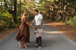 A family of three holds hands on a forest path, with parents guiding their toddler between tall trees in warm sunlight.