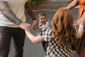 A joyful toddler boy holds hands with tattooed dad and redhead sister during a warm family outdoor photo session.