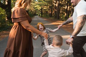 Family of four joyfully spinning children on a scenic tree-lined path in warm afternoon sunlight.