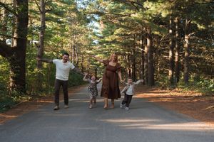 Family of four joyfully jumping on a tree-lined road surrounded by tall pine trees in a forest setting.