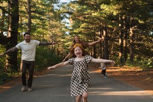 Family joyfully running on a forest path with arms outstretched, surrounded by tall pine trees in warm golden light.