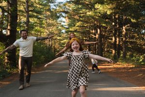 A joyful red-haired girl runs with arms out on a forest path as her family plays behind her.