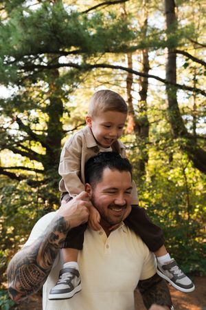 Smiling tattooed father carrying young boy on shoulders in a sunny forest setting.