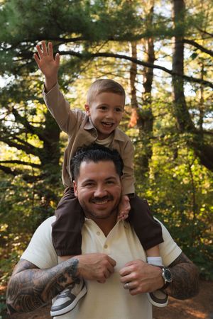 A smiling father with tattoos carries his happy young son on his shoulders in a sunlit forest setting.