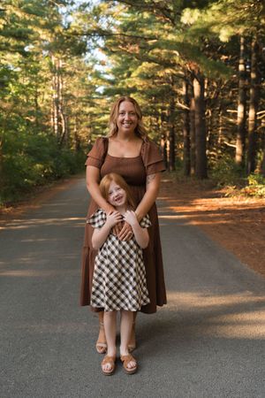 Mother in brown dress hugging red-haired daughter in plaid dress on a forest road.