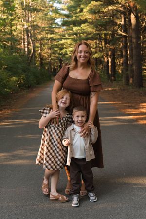 Mother in brown dress smiling with two young children on a tree-lined road in autumn.