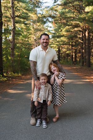A tattooed father smiles with his two young children on a sunlit tree-lined road outdoors.