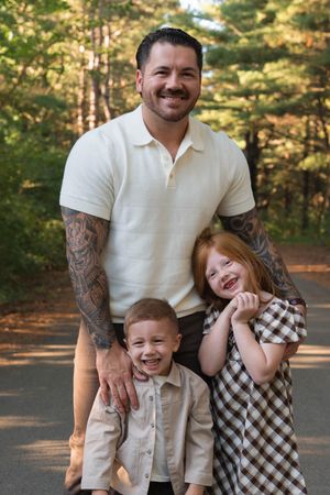 Tattooed father smiles proudly with two young redheaded children on a tree-lined path outdoors.