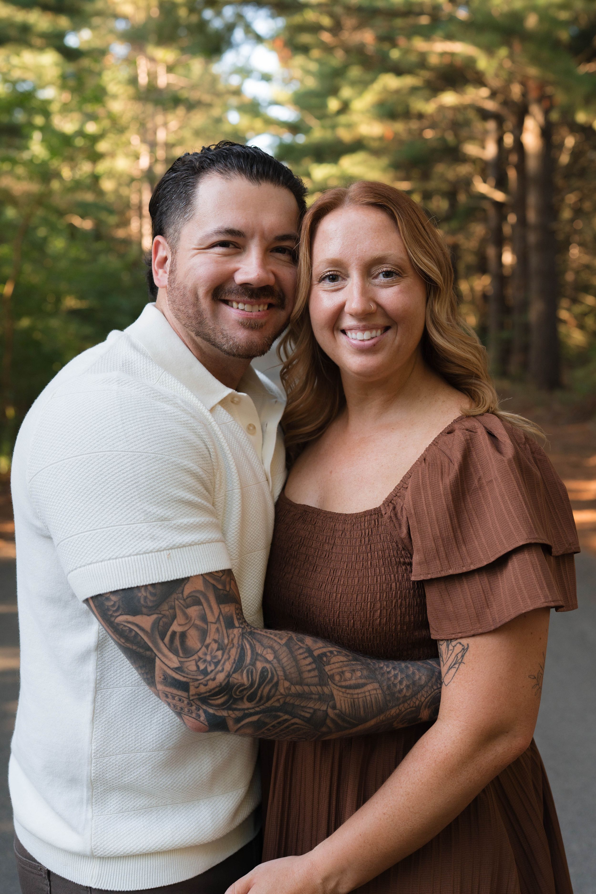 A smiling couple embraces outdoors, the man with tattooed arms wearing a cream polo beside a woman in brown dress.