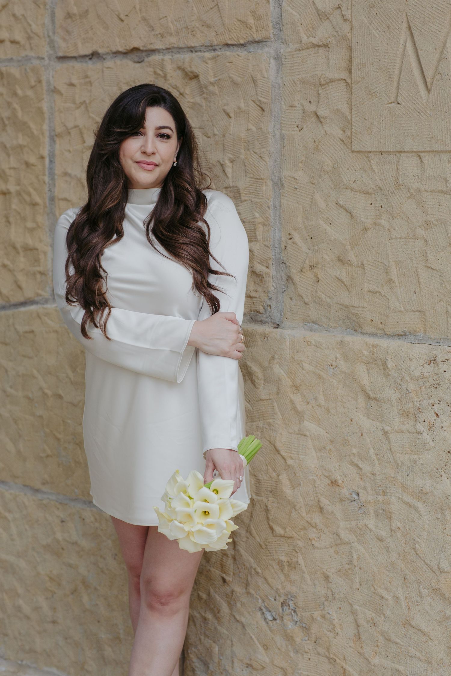 Young woman in white mini dress holding white tulip bouquet, posing against a stone wall with long wavy dark hair.