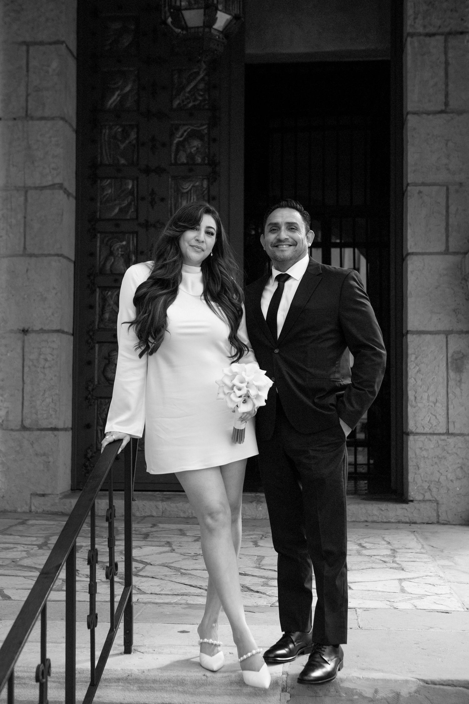 Black and white photo of a bride in short white dress holding bouquet with groom in suit on building steps.