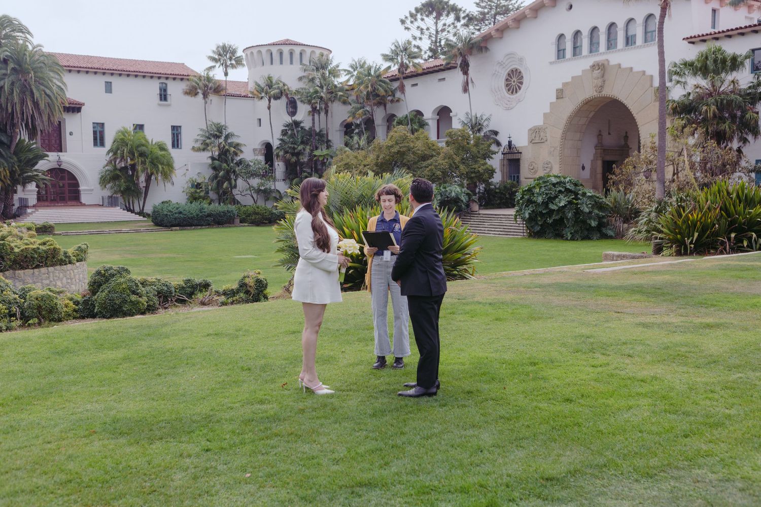 Three people gather on green lawn before grand Spanish Colonial Revival building with arched entrance and palm trees.