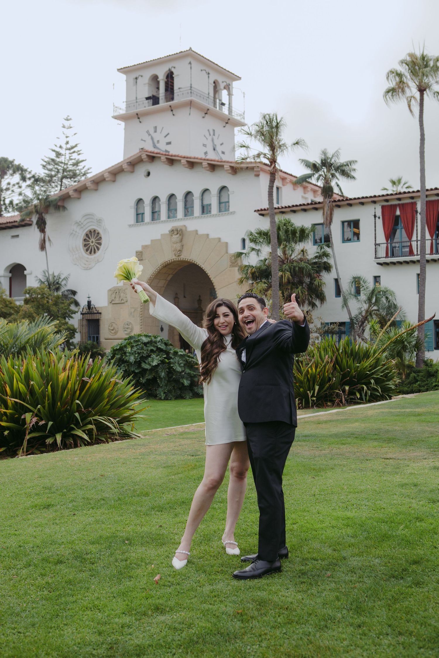 Joyful couple celebrating their wedding outside Santa Barbara Courthouse, bride in white mini dress holding bouquet.