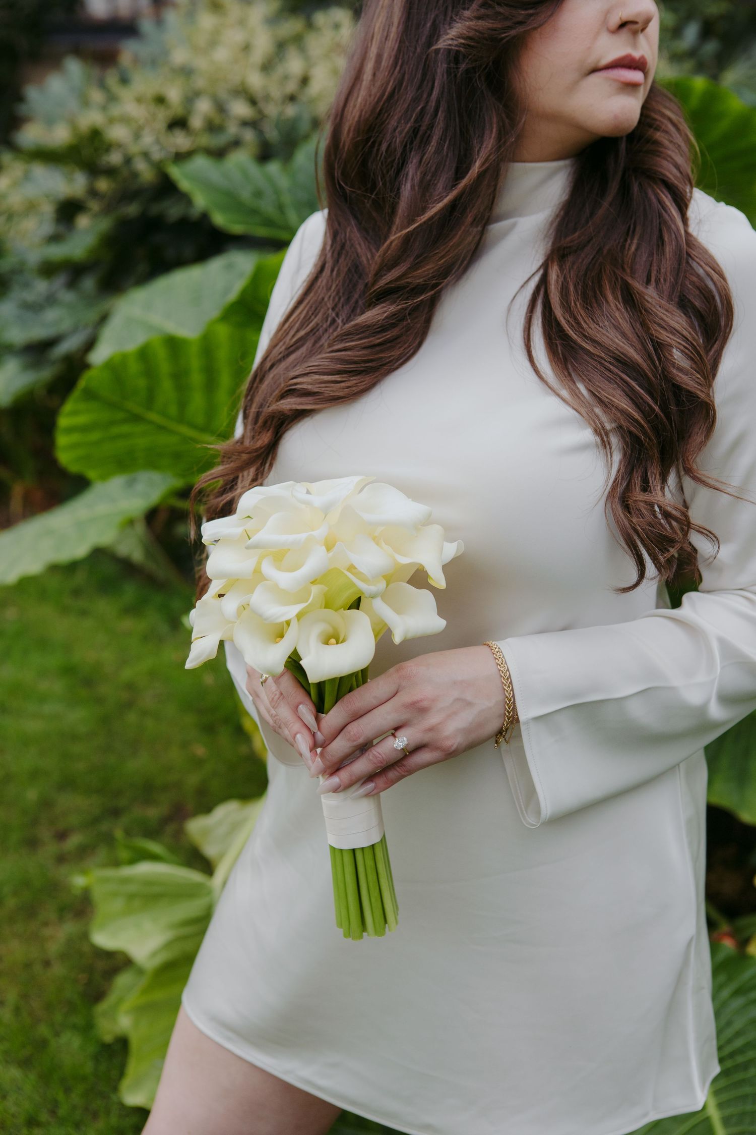 Bride in white long-sleeve mini dress holding elegant white calla lily bouquet in lush garden setting.