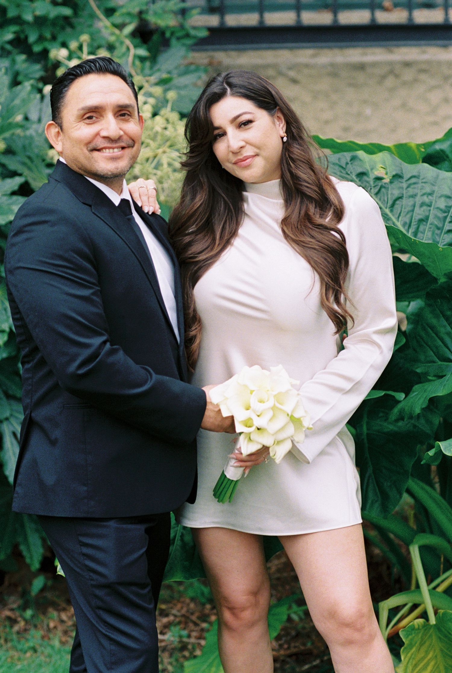 Couple posing outdoors, man in black tuxedo and woman in white mini dress holding calla lily bouquet, surrounded by lush greenery.