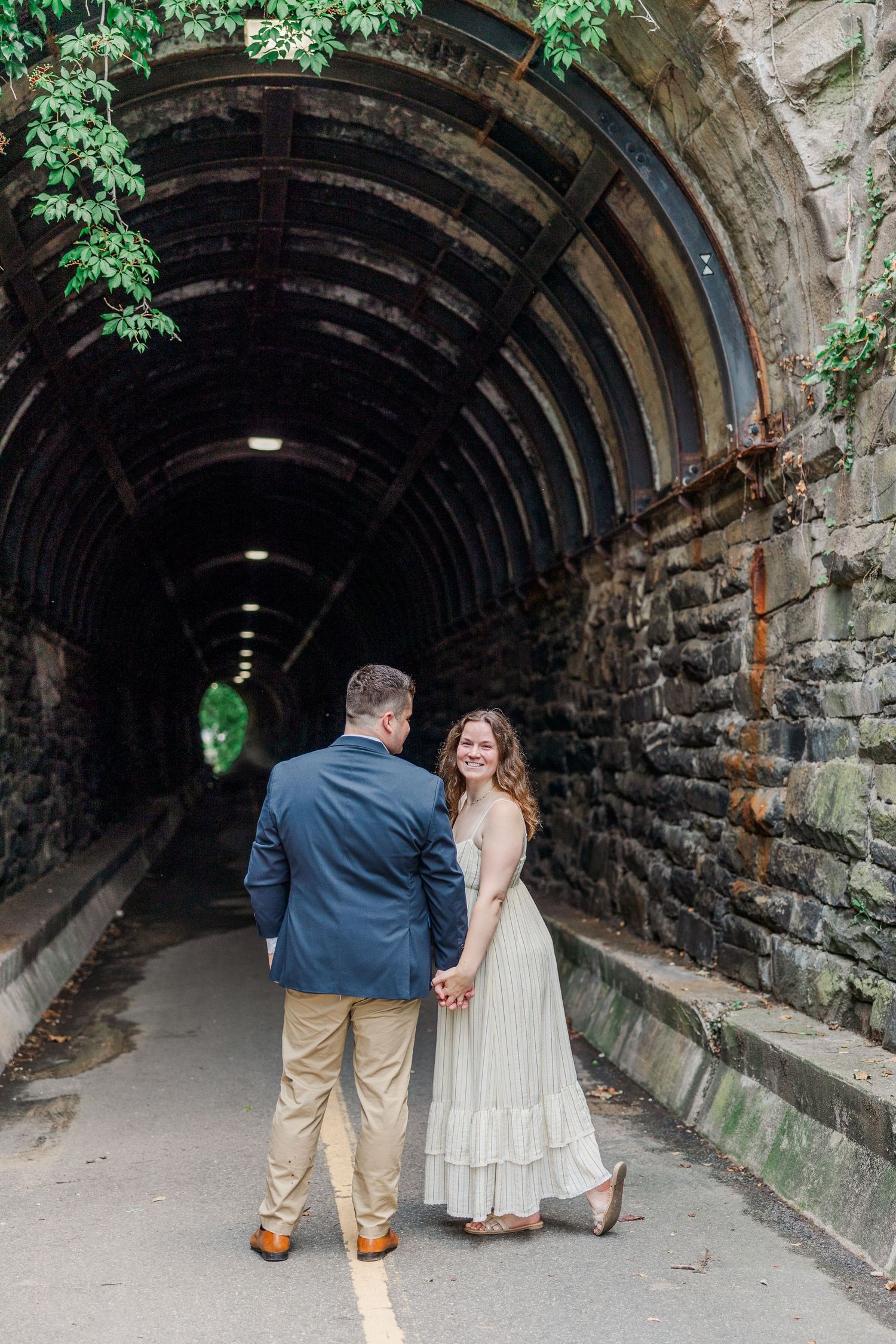 Smiling couple holding hands at entrance of stone tunnel archway surrounded by green foliage.