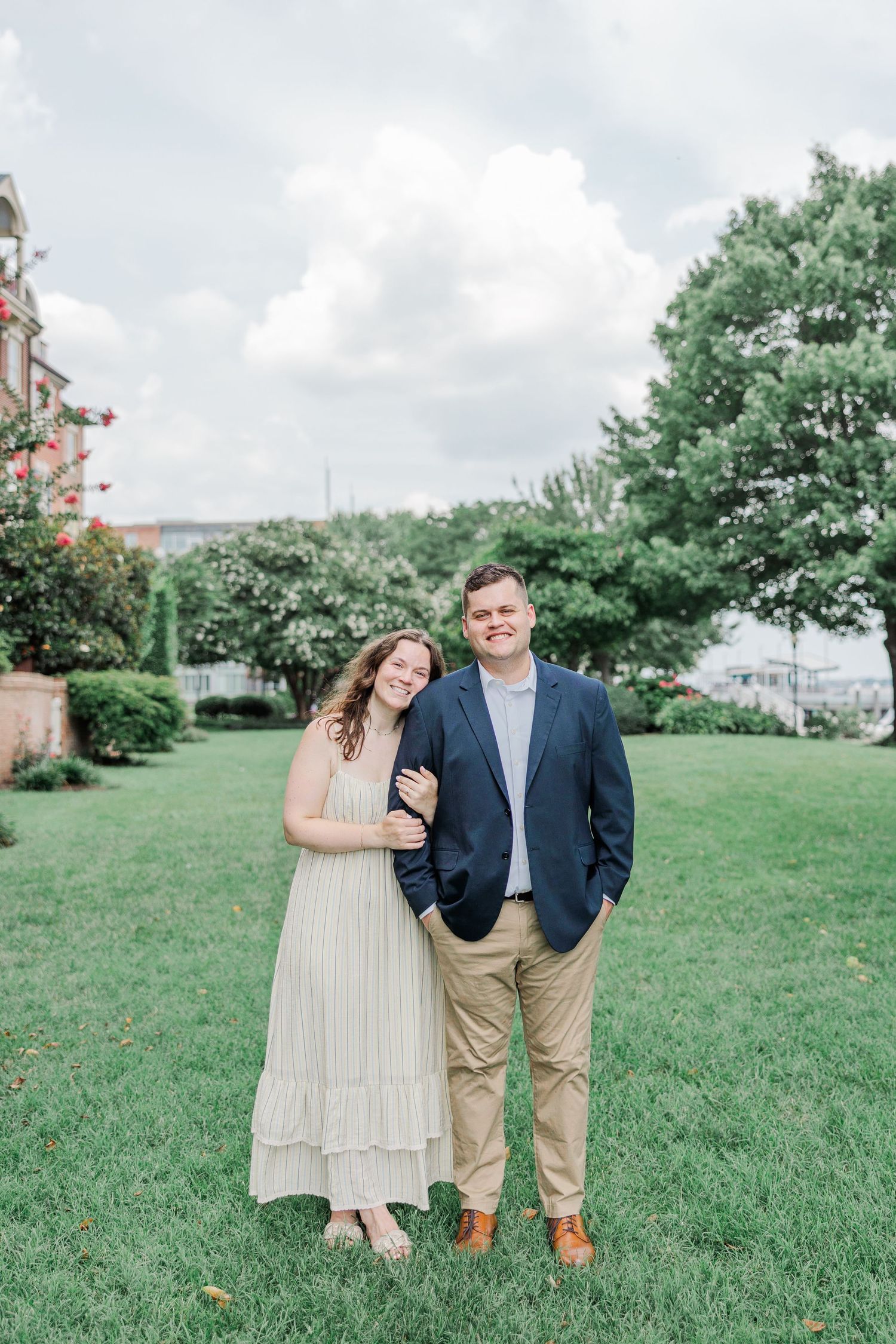 A couple smiles together on a green lawn, woman in cream dress, man in navy blazer and khakis, lush trees behind them.